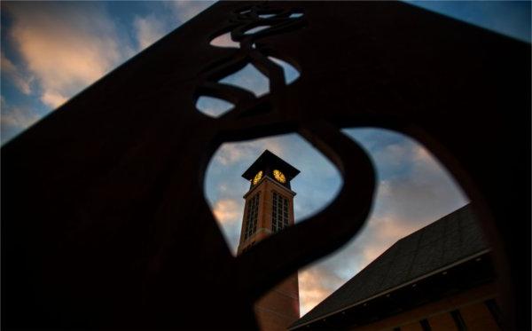 A carillon tower is seen through the shapes of a metal sculpture during sunrise. 